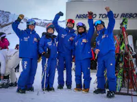 Five people in matching blue ski suits and helmets stand on snow, smiling and raising their arms in celebration in front of a ski resort building. Ski equipment is visible beside them.