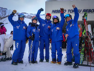 Five people in matching blue ski suits and helmets stand on snow, smiling and raising their arms in celebration in front of a ski resort building. Ski equipment is visible beside them.