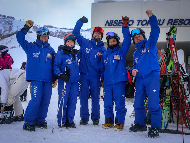 Five people in matching blue ski suits and helmets stand on snow, smiling and raising their arms in celebration in front of a ski resort building. Ski equipment is visible beside them.
