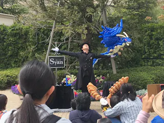 A street performer in a black suit and top hat dramatically poses while holding a large blue balloon sculpture of a dragon, entertaining a crowd outdoors. Several audience members have balloon animals on their heads or arms.