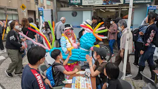 A colorful clown with a balloon costume entertains a group of children and adults seated at tables outdoors, while people wearing masks walk by on a busy street.