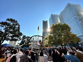A crowd gathers outdoors near a tall play structure and trees, with modern high-rise buildings in the background under a clear blue sky and bright sunlight.