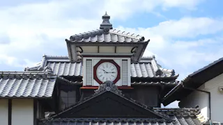 Traditional Japanese building with a tiled roof and a large round clock centered on the upper facade; the sky is partly cloudy in the background.