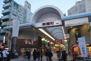 A bustling Japanese shopping street with a large arched entrance reading "1st," lined with shops, signs, and people walking and cycling under a covered arcade at dusk.