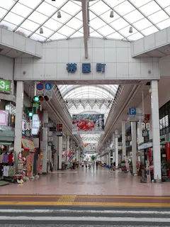 A covered shopping street in Japan with shops on both sides, signs in Japanese, bright lighting from a glass roof, and a few people walking and cycling along the wide pedestrian path.