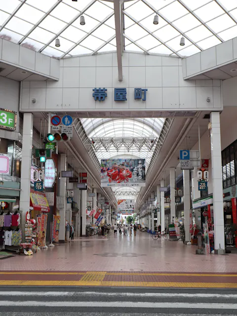 A covered shopping street in Japan with shops on both sides, signs in Japanese, bright lighting from a glass roof, and a few people walking and cycling along the wide pedestrian path.