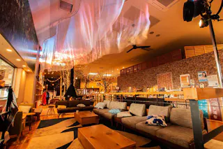 A cozy, warmly lit lounge with modern sofas, wooden tables, brick walls, and decorative lights. A translucent fabric is draped across the ceiling, creating a soft, artistic ambiance.