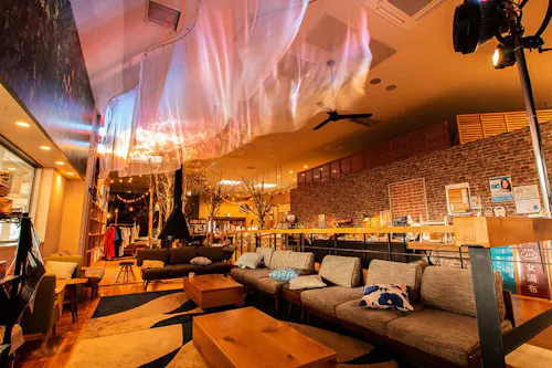 A cozy, warmly lit lounge with modern sofas, wooden tables, brick walls, and decorative lights. A translucent fabric is draped across the ceiling, creating a soft, artistic ambiance.