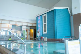 Indoor bathhouse with a turquoise sauna hut beside a large hot tub. Neon “SAUNA” sign glows above the hut’s door. Walls are tiled, with sinks and mirrors in the background. Bright, clean, and inviting atmosphere.