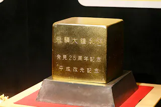 A gold-colored cubical object with Japanese inscriptions on its sides, displayed on a black and red pedestal against a dark background.