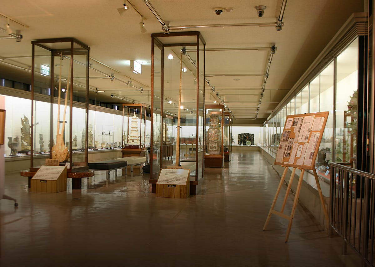 A spacious museum gallery with glass display cases containing various artifacts, wide walkways, informational signs, and a bulletin board with papers and images near the entrance. The area is well-lit with overhead lights.