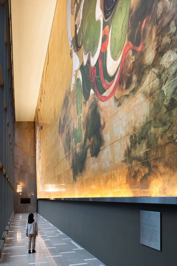 A person walks down a modern, spacious hallway with high ceilings, beside a large, colorful mural featuring abstract designs and swirling patterns on the wall. Warm lights illuminate the mural from below.