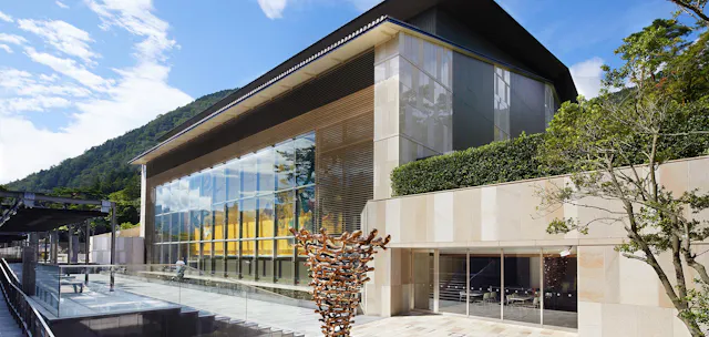 A modern building with large glass windows and stone walls, surrounded by greenery and mountains. In the foreground is a unique outdoor sculpture made of interwoven, bronze-colored branches. The sky is bright and partly cloudy.