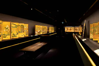 A dimly lit museum gallery displays illuminated Japanese folding screens featuring intricate gold and colorful nature scenes along dark walls, with benches and display cases in the center.