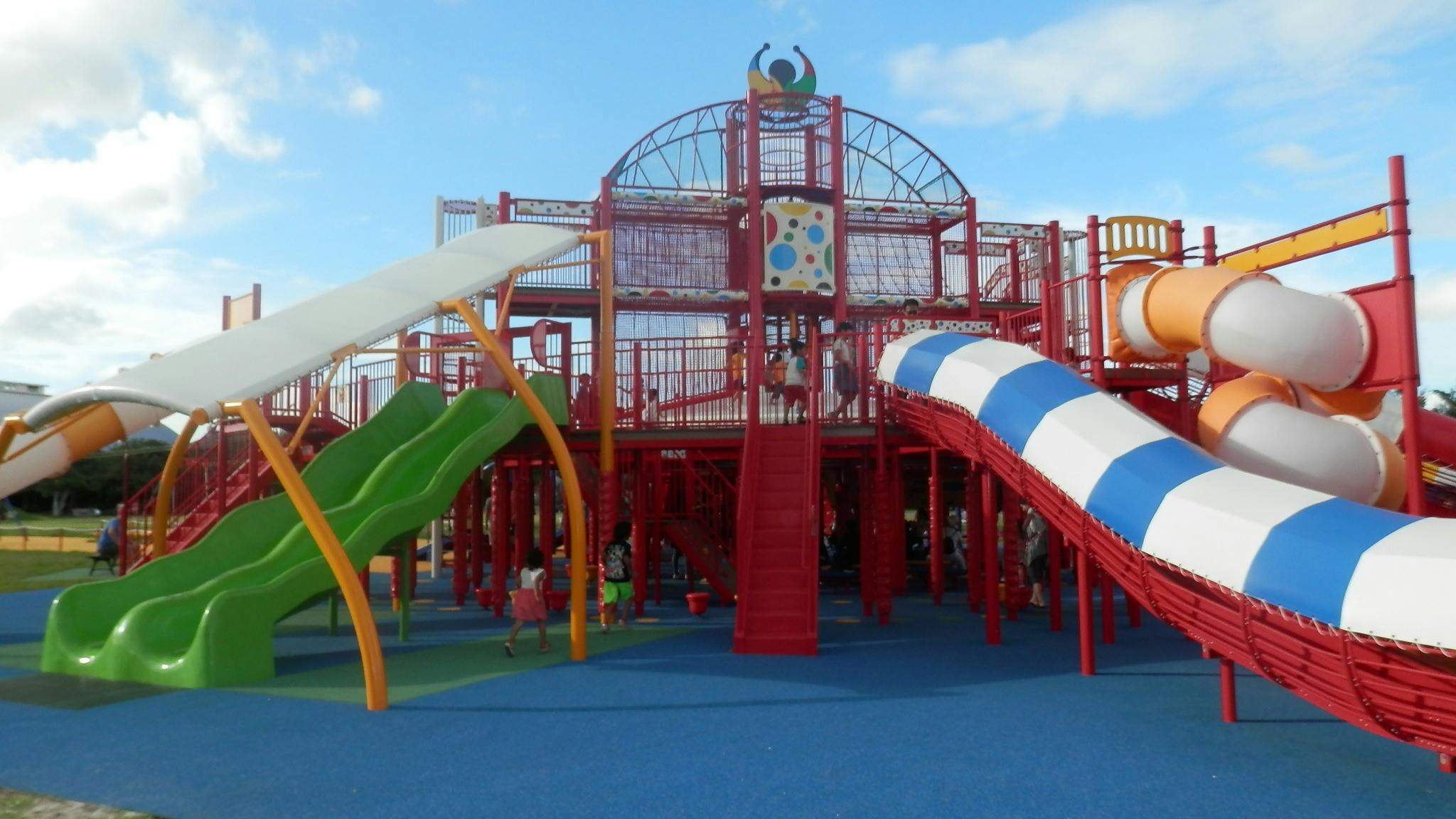 A colorful playground with red climbing structures, green slides, a blue and white striped slide, an orange spiral tube slide, and a blue safety mat under a partly cloudy sky.