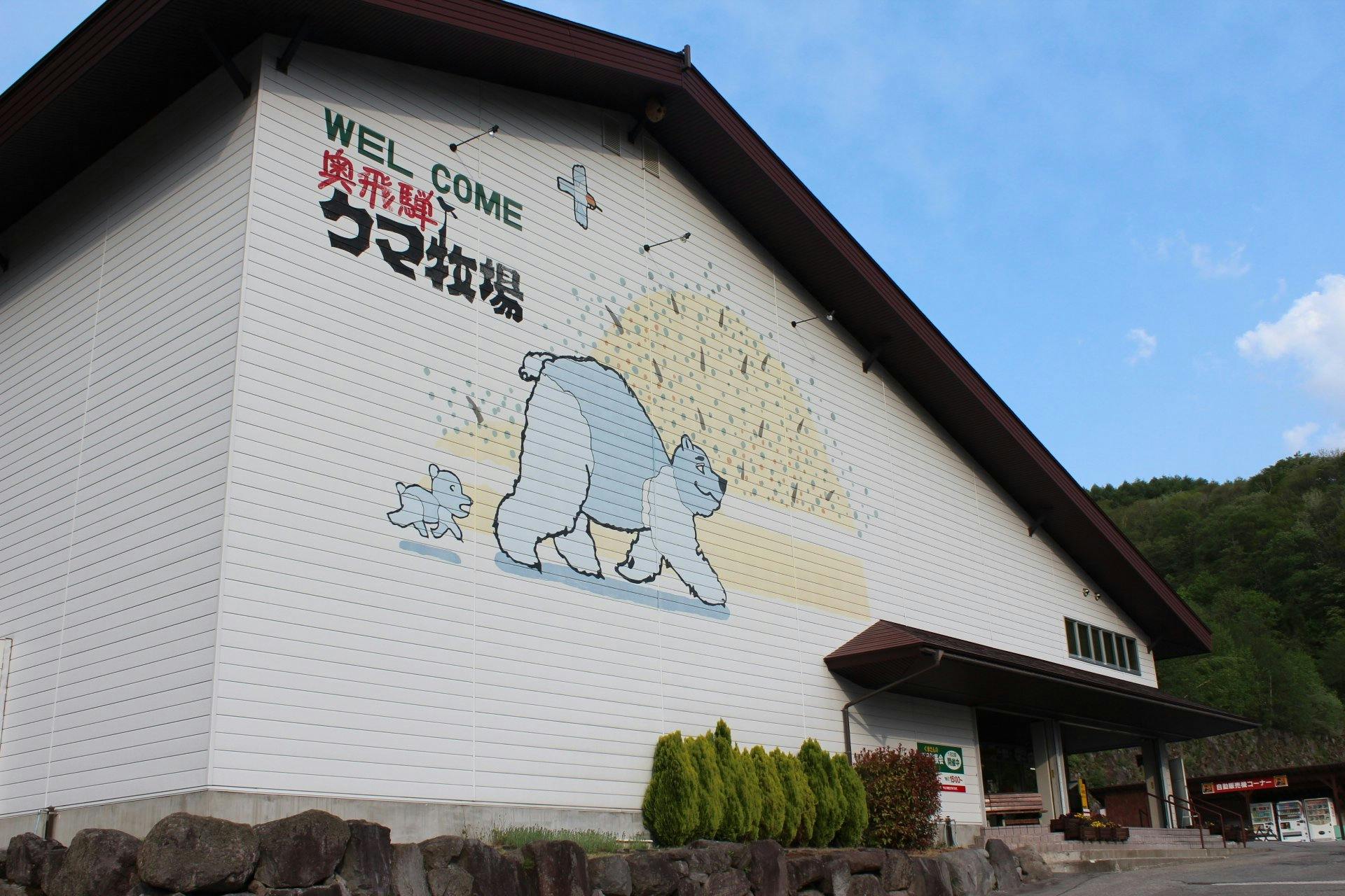 A large white building with a mural of two cartoon sheep facing a haystack is shown. Japanese text and the word "WELCOME" are painted above the mural. Green shrubs line the building and trees are visible in the background.
