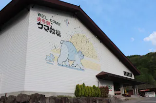 A large white building with a mural of two cartoon sheep facing a haystack is shown. Japanese text and the word "WELCOME" are painted above the mural. Green shrubs line the building and trees are visible in the background.