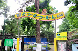 Entrance to a zoo with a colorful sign in Japanese and English reading "Omia Park Zoo." Cartoon animals decorate the sign, and trees and fences are visible in the background.