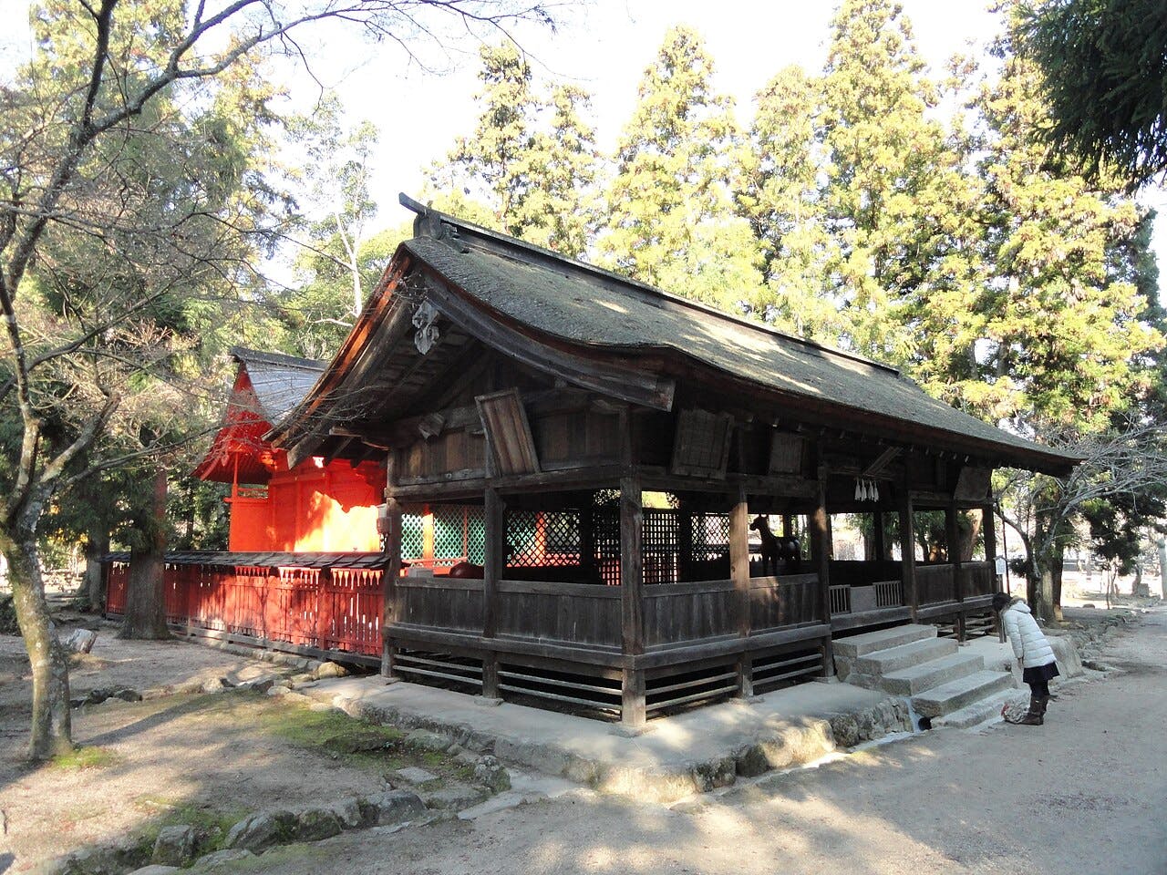 A traditional wooden Shinto shrine building with a sloped roof, surrounded by trees. A person stands near the entrance, and a smaller red shrine structure is visible in the background.