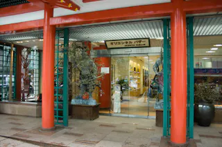 A storefront with large red columns, glass windows, and statues flanking the entrance. The sign above the door reads "オリエンタル バザー" (Oriental Bazaar). The interior displays various goods and decorations.