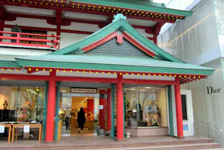 A store entrance with a green and red traditional Japanese-style roof, labeled "Oriental Bazaar," next to a modern Dior storefront. People can be seen entering and exiting the building.