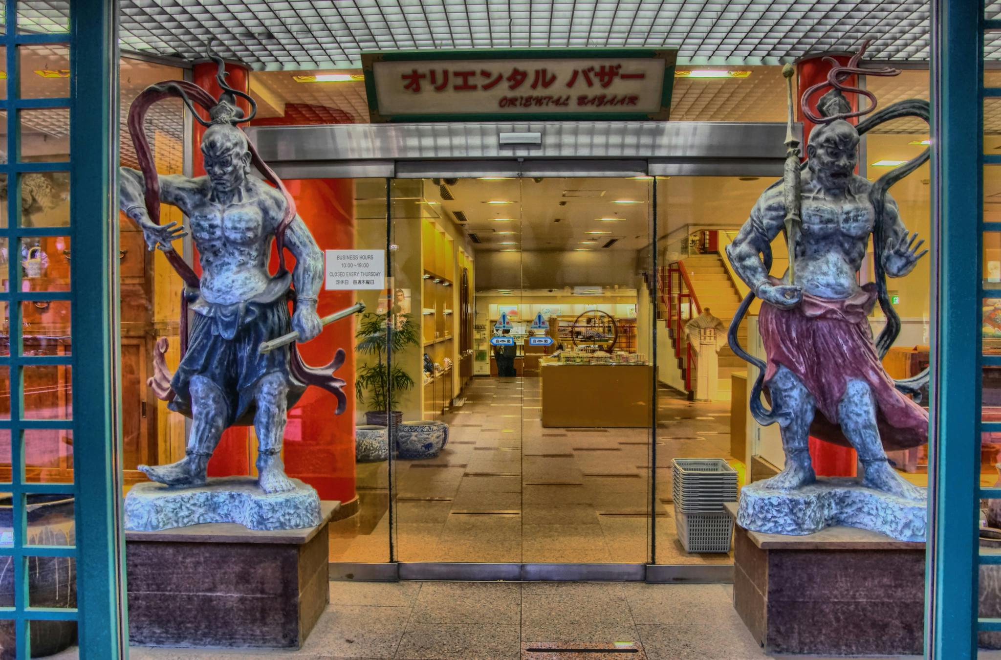 Two large warrior statues stand on either side of a glass entrance to a store called "Oriental Bazaar" with Japanese writing above the door; the interior is brightly lit and filled with various items.