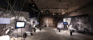 A modern museum exhibit features large black-and-white photos on the walls, video screens on stands, scattered debris, and dramatic lighting, creating an immersive and somber atmosphere.