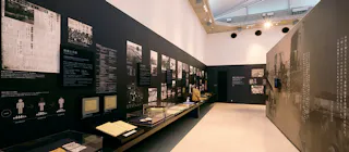 A museum exhibit with black walls displaying historical photos, documents, and newspaper clippings in glass cases and on panels. The space is well-lit with modern lighting and a high ceiling.