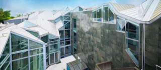 A modern building with angular, geometric architecture featuring large glass windows, metal roofing, and green-tinted wall panels under a clear sky.