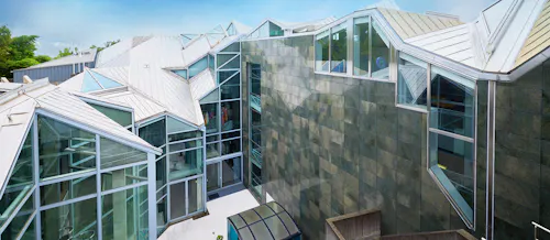 A modern building with angular, geometric architecture featuring large glass windows, metal roofing, and green-tinted wall panels under a clear sky.