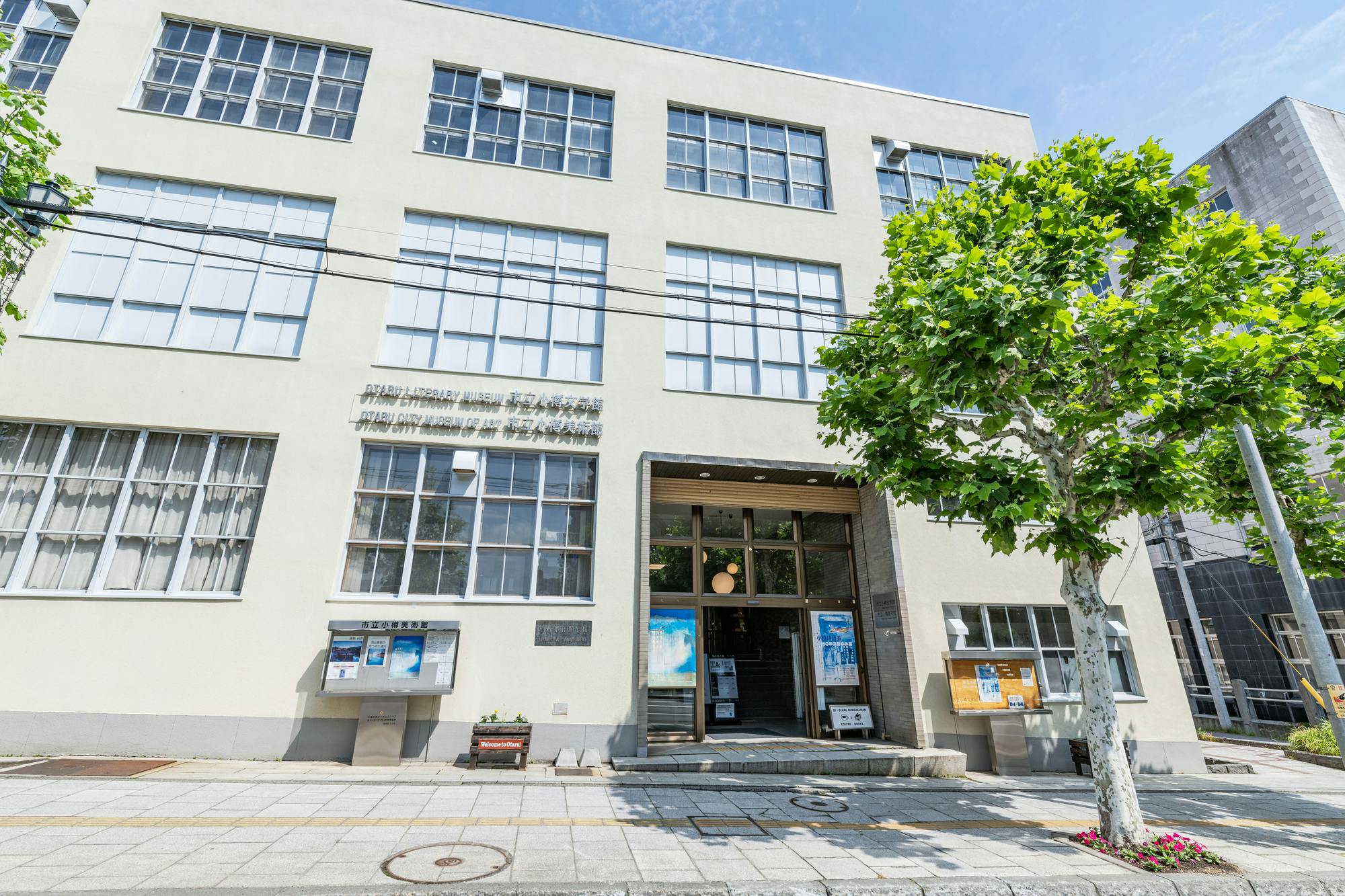 A three-story building with large windows and a main entrance under a canopy. Posters are displayed near the door, and a leafy tree stands on the sidewalk in front of the building on a sunny day.