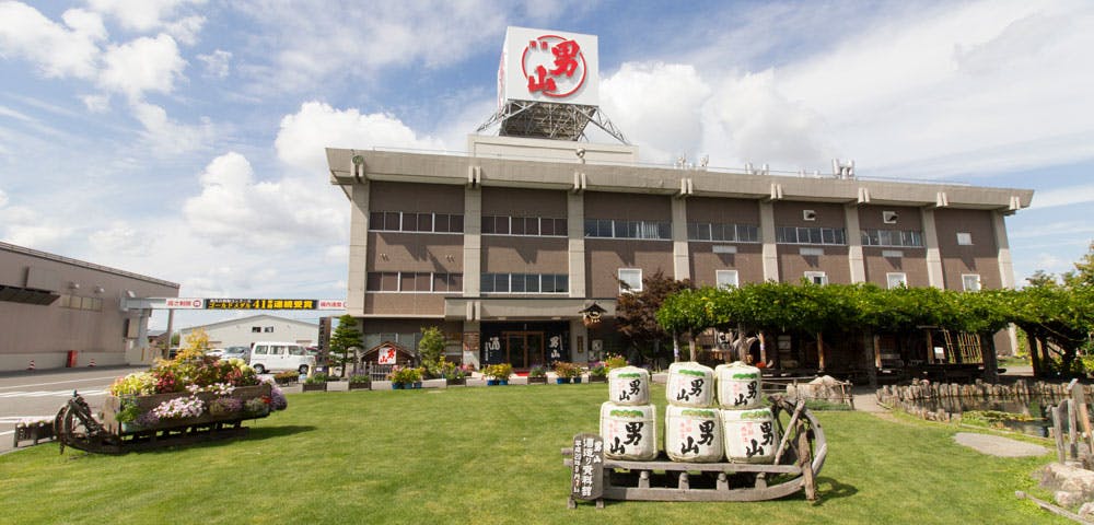 A large building with a red sign on the roof stands behind a well-kept lawn decorated with flower beds and stacked sake barrels under a blue sky with clouds.