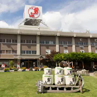 Otokoyama Sake Brewery A large building with a red sign on the roof stands behind a well-kept lawn decorated with flower beds and stacked sake barrels under a blue sky with clouds.