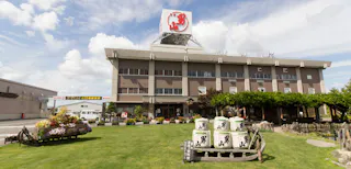 A large building with a red sign on the roof stands behind a well-kept lawn decorated with flower beds and stacked sake barrels under a blue sky with clouds.