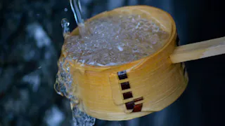 A wooden ladle is being filled with clear water, causing some water to overflow down its side. The background is blurred, highlighting the motion and texture of the water.