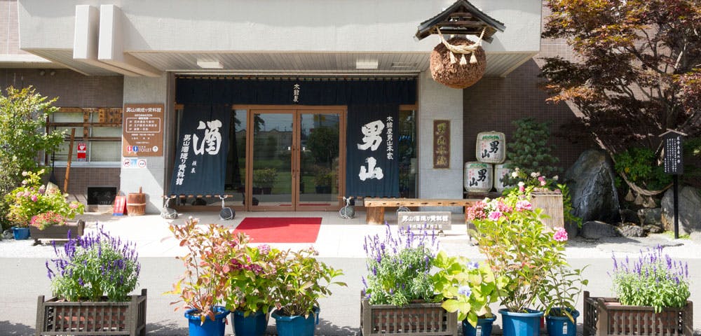 Entrance of a Japanese sake brewery with noren curtains, potted plants, and a sugidama (cedar ball) hanging above the doorway. Japanese signage and banners are displayed around the entrance.