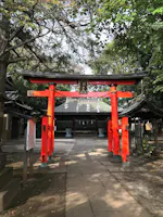A bright red torii gate stands at the entrance of a Shinto shrine, surrounded by trees and traditional buildings, with sunlight filtering through the leaves. The path leads to the shrine’s main structure in the background.