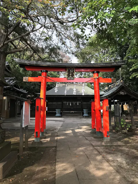 A bright red torii gate stands at the entrance of a Shinto shrine, surrounded by trees and traditional buildings, with sunlight filtering through the leaves. The path leads to the shrine’s main structure in the background.