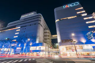 A night view of a city intersection with modern white buildings, including the Creston Hotel and Parco, both illuminated with blue and white lights. Light trails from cars and bare trees are visible in the foreground.