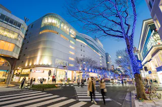 A lively city street at dusk with people crossing a crosswalk, modern buildings, PARCO department store, and trees decorated with blue lights. The scene feels energetic and festive.