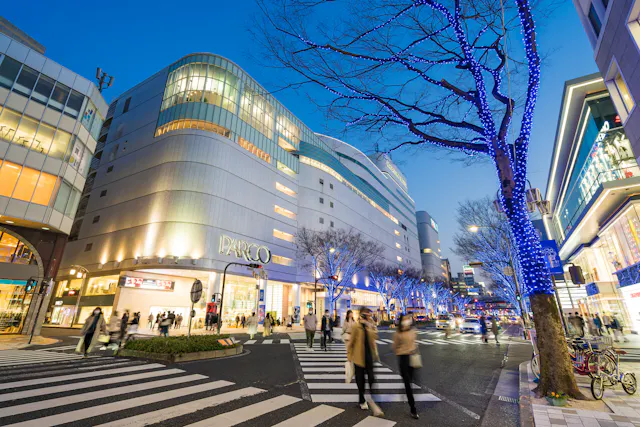 A lively city street at dusk with people crossing a crosswalk, modern buildings, PARCO department store, and trees decorated with blue lights. The scene feels energetic and festive.