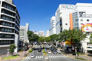 A busy city street with cars and pedestrians, lined with tall buildings including one marked "PARCO" and another featuring a large cartoon animal face. Trees line the sidewalks on a bright, clear day.