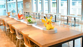 A spacious, bright café with wooden tables and chairs, featuring large Pokémon character figures, including Pikachu, displayed as table centerpieces. Large windows provide ample natural light.