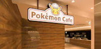 A brightly lit interior of the Pokémon Cafe, featuring a wooden wall and a large sign with Pikachu wearing a chef’s hat above the entrance. The kitchen area and food counter are visible in the background.