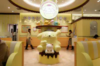 A themed café interior featuring a large Pompompurin character statue in the center, surrounded by pastel-colored decorations, plush toys, and people in matching uniforms walking by.
