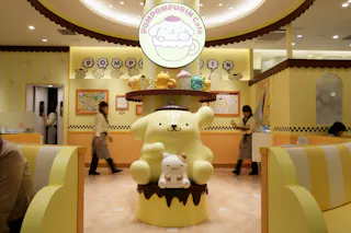 A themed café interior featuring a large Pompompurin character statue in the center, surrounded by pastel-colored decorations, plush toys, and people in matching uniforms walking by.