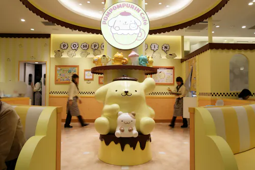 A themed café interior featuring a large Pompompurin character statue in the center, surrounded by pastel-colored decorations, plush toys, and people in matching uniforms walking by.