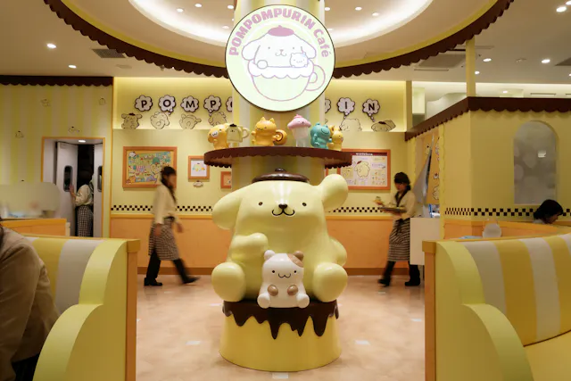 A themed café interior featuring a large Pompompurin character statue in the center, surrounded by pastel-colored decorations, plush toys, and people in matching uniforms walking by.