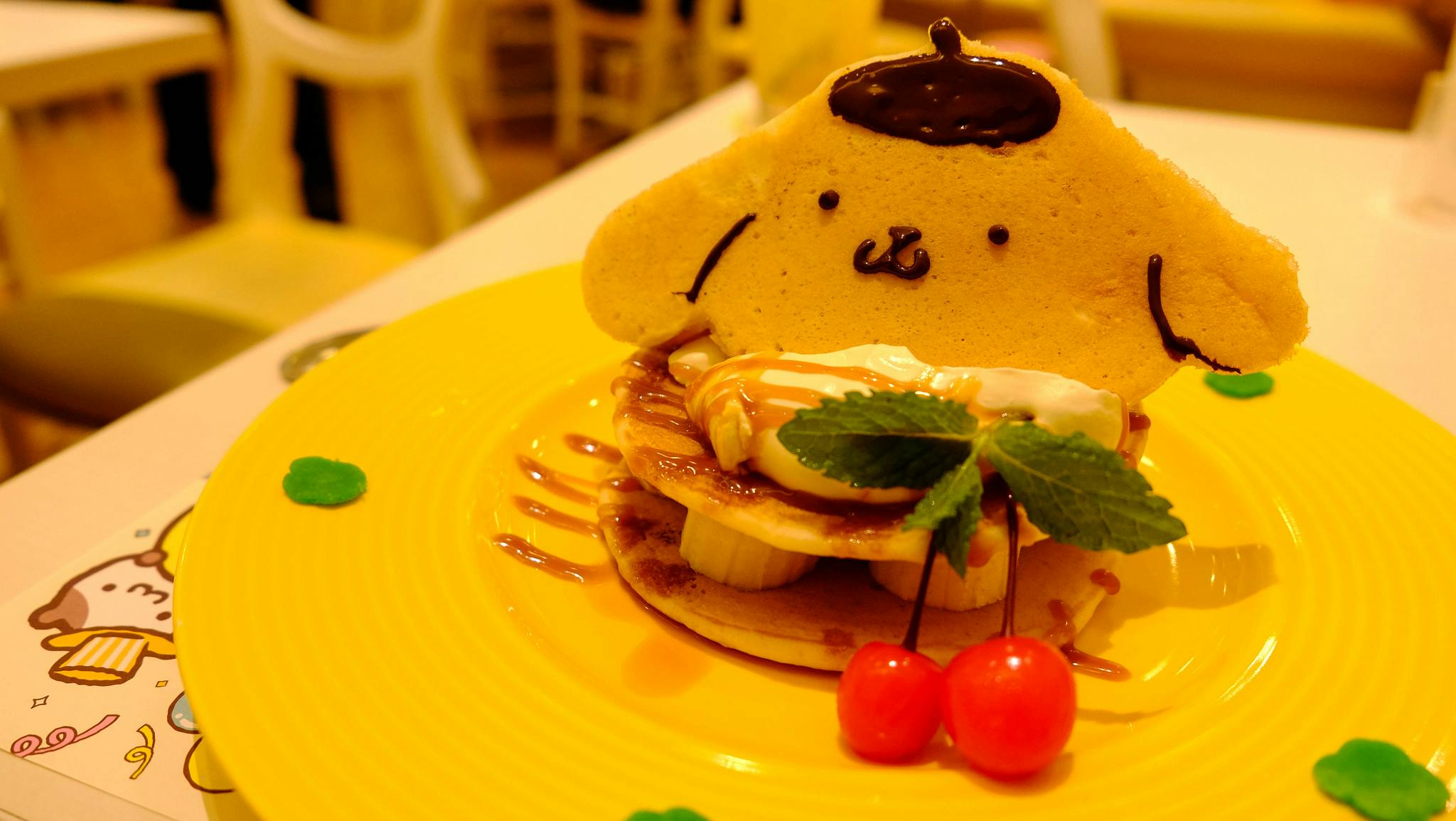 A plate with a cute dessert: pancakes topped with banana slices, cream, and caramel, decorated with a character-shaped cookie, two cherries, and a mint leaf, on a yellow plate in a brightly lit café.