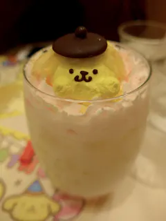 A glass filled with a creamy dessert topped with a yellow character resembling a dog, made of whipped cream or mousse, wearing a brown beret. The dessert is placed on a colorful, cartoon-themed placemat.